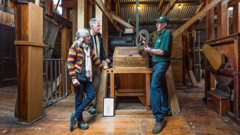 A volunteer showing a man and woman some of the mill machinery