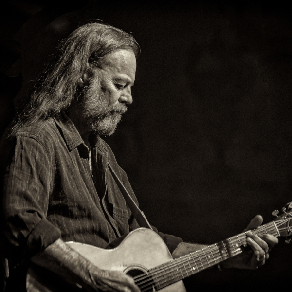 Black and white profile image of a man with long hair and a beard playing a guitar.