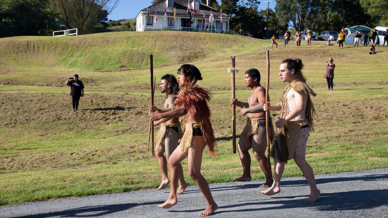 A group of young Māori men welcome guests during a pōwhiri with Māngungu Mission house in the background