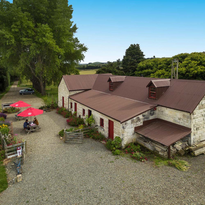 Aerial view of the main Totara Estate building made of Ōamaru stone, picnic tables with red umbrellas and people sitting beneath them