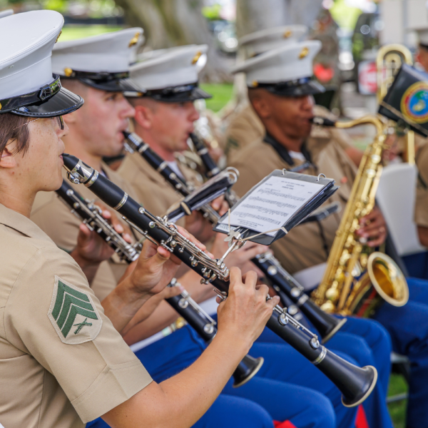 Uniformed men and women in tan shirts and blue trousers play clarinets and saxophone outdoors.