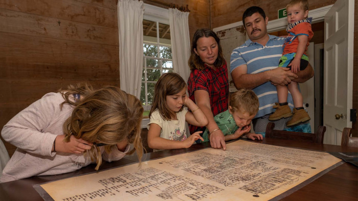 Mother, Father and four children view a copy of the Treaty of Waitangi on the table in the mission house