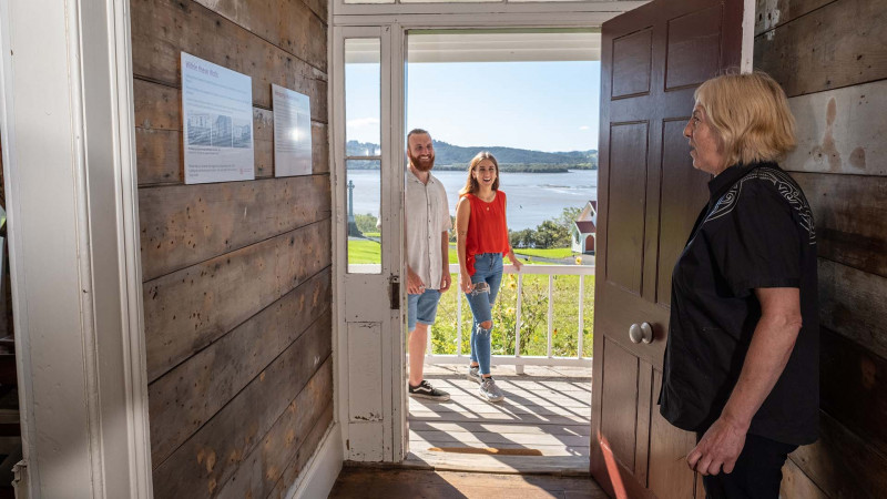 Staff member standing inside the mission house welcomes a young couple into the house with the view of the Hokianga Harbour in the background