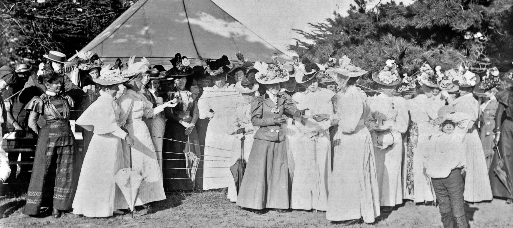 A group of women in hats and dresses gather in front of a marquee. 