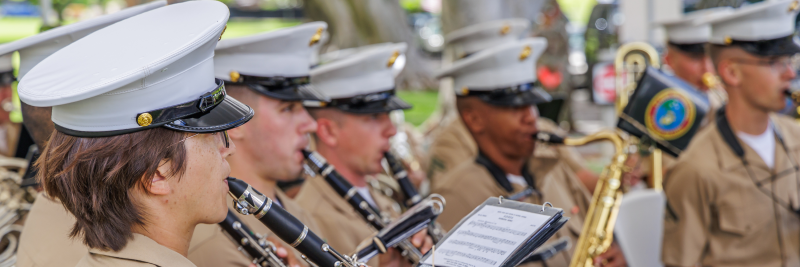 Uniformed men and women in tan shirts and hats play clarinets and saxophone outdoors.